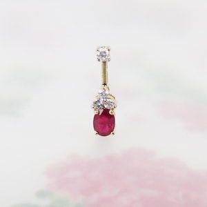 May include: A gold-toned earring featuring a vibrant red oval-cut gemstone at the base, accented with three small white gemstones above. A single white gemstone tops the earring. The jewelry is set against a soft, floral background.