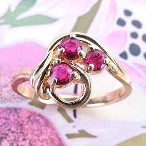 May include: A gold ring featuring three round, vibrant pinkish-red gemstones set in a swirling, polished gold band. The design showcases a bypass-style setting where the metal curves around the stones, creating an open, fluid, and continuous loop.
