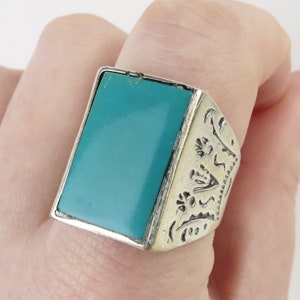 May include: A silver ring featuring a rectangular turquoise stone. The ring's sides are decorated with etched patterns. The turquoise stone is a vibrant, solid colour. The ring is worn on a finger.