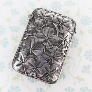 May include: A rectangular silver-toned metal case featuring a detailed embossed pattern of three-leaf clovers across the entire front surface. The metal has a darkened, oxidised finish in the recessed areas, highlighting the raised clover leaves.