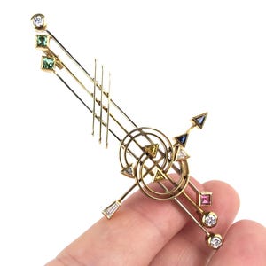 May include: A gold-toned brooch with geometric shapes and colorful gemstones. The brooch features a series of lines, circles, and arrows, adorned with diamonds, emeralds, sapphires, and a pink gemstone. The design is intricate and artistic.