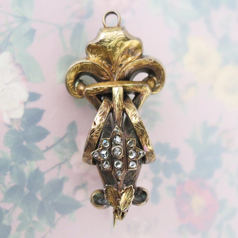 May include: Antique gold-toned pendant with a fleur-de-lis design. The pendant features intricate detailing, including a starburst of clear stones and floral accents. The piece has a pin on the back and a loop at the top for wear.