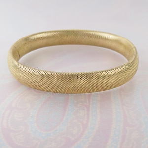 May include: A gold-toned bangle bracelet featuring a fine, cross-hatched textured surface throughout the entire exterior. The circular jewelry piece has a rounded profile and a consistent width, showcasing a metallic finish with a subtle sheen.