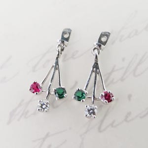 May include: A pair of silver earrings with a unique design. Each earring features a triangular arrangement with a red, green, and clear gemstone. The earrings have a post-back closure.