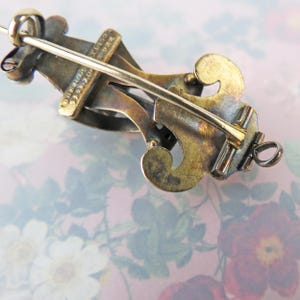 May include: Close-up of a vintage brooch with a decorative design. The brooch features a gold-toned metal with intricate details and a pin closure. The design includes curved elements and a bar across the center.