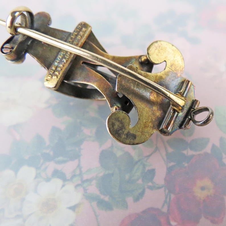 May include: Close-up of a vintage brooch with a decorative design. The brooch features a gold-toned metal with intricate details and a pin closure. The design includes curved elements and a bar across the center.