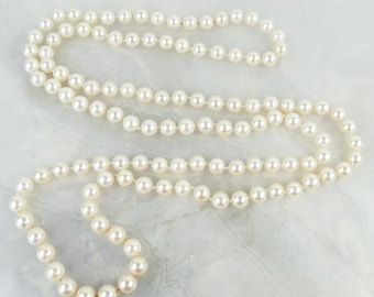 May include: A long necklace of creamy white pearls. The pearls are round and uniform in size, creating a classic and timeless design. The necklace is displayed on a light-coloured surface, accentuating the pearls' lustre.