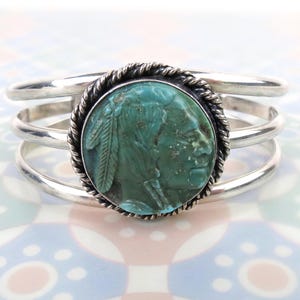 May include: Sterling silver cuff bracelet featuring a turquoise-colored stone with a carved Native American profile. The stone is encircled by a twisted rope design and set within a silver frame. The bracelet has three parallel silver bands.