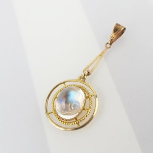 May include: A gold pendant with a large, oval, iridescent moonstone. The moonstone is set within a circular frame with a beaded edge and a delicate gold bale. The pendant hangs from a slender, triangular gold chain.