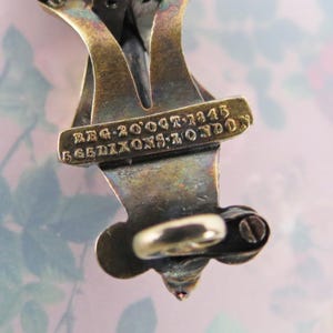 May include: Close-up of a vintage metal jewelry piece with a tarnished, aged appearance. The piece has an intricate design with a small, round clasp and a decorative element at the bottom. Text on the piece reads "REG 20'OCT'1343" and "565 DIXONS: LONDON".