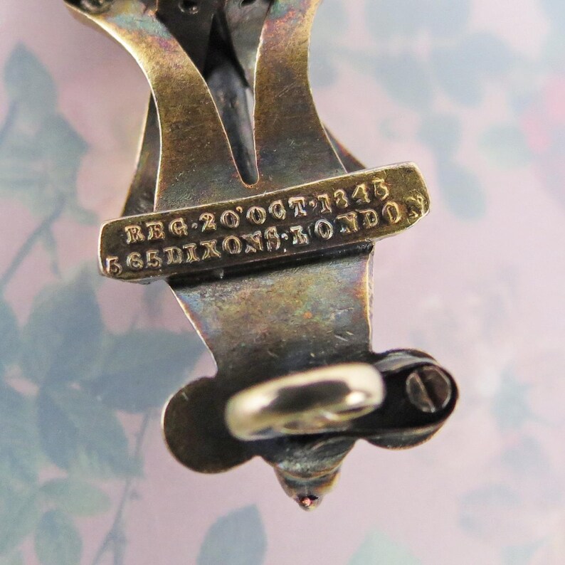 May include: Close-up of a vintage metal jewelry piece with a tarnished, aged appearance. The piece has an intricate design with a small, round clasp and a decorative element at the bottom. Text on the piece reads "REG 20'OCT'1343" and "565 DIXONS: LONDON".