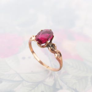 May include: A gold ring featuring a single oval-cut deep pink gemstone held in a prong setting. The band is crafted from polished gold and displays a decorative, swirling metalwork design on both sides of the central stone setting.