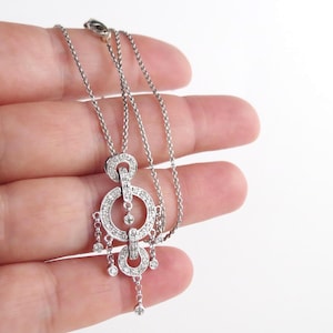 May include: A silver-toned necklace featuring a pendant with two circular, crystal-encrusted rings connected vertically. Several small, dangling chains with individual round crystals hang from the rings, creating a layered, chandelier-like appearance.