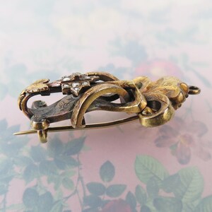 May include: An antique gold-toned brooch with intricate floral and scrollwork designs. The brooch features a central cluster of small, sparkling stones and a pin closure on the back. The metal has a tarnished appearance, adding to its vintage charm.