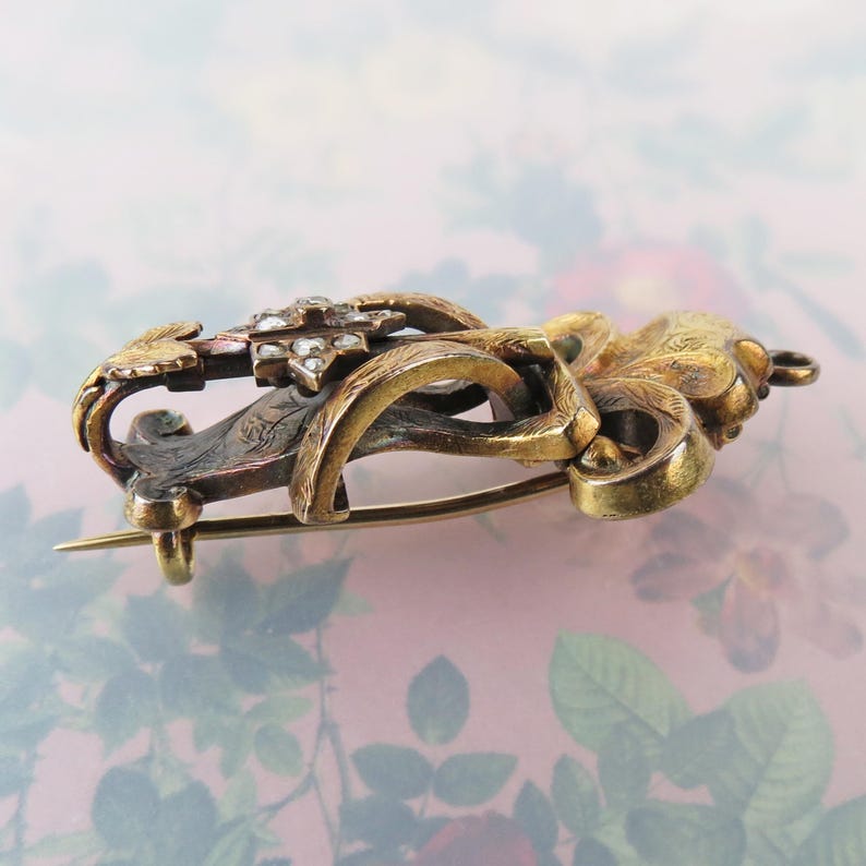 May include: An antique gold-toned brooch with intricate floral and scrollwork designs. The brooch features a central cluster of small, sparkling stones and a pin closure on the back. The metal has a tarnished appearance, adding to its vintage charm.