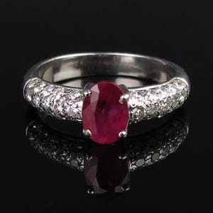 May include: A silver-toned ring featuring a central oval-cut deep red ruby gemstone held by four prongs. The band is encrusted with numerous small, clear, sparkling stones along the sides, creating a bright contrast against the dark red center.