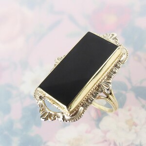 May include: A rectangular ring featuring a smooth, flat black onyx stone set within a gold bezel. The setting is framed by an intricate, decorative silver-toned metal border with ornate scrollwork details and a thin, polished gold band.