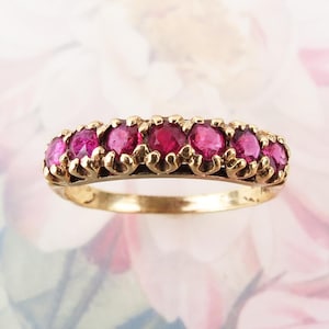 May include: A gold band ring featuring seven round, deep pinkish-red gemstones set in a row. Each stone is held in place by small, individual metal prongs, creating a continuous line of color across the top of the polished gold band.