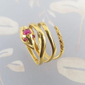 May include: A gold snake ring featuring a coiled band that wraps around the finger. The snake head is set with three small, round, vibrant pink rubies representing the eyes and forehead. The tail end of the band has a textured, cross-hatched pattern.