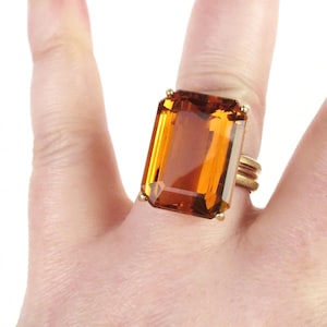 May include: A large rectangular faceted amber-coloured gemstone ring set in a gold-toned metal band. The stone features a prominent emerald cut with clear transparency and warm orange-brown hues, secured by four small prongs on a simple band.