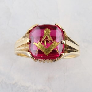 May include: A gold-colored ring with a large, square, ruby-red stone. The stone features a gold-colored Masonic symbol. The ring has a classic design with a thick band and decorative shoulders. A vintage piece.