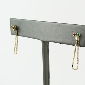May include: A pair of gold drop earrings featuring a small cluster of a round red stone, a round green stone, and a clear white stone at the top, connected to a delicate gold box chain that loops down to create a slender, dangling silhouette.