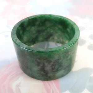 May include: A solid green jade ring with a cylindrical shape. The ring has a polished surface with dark green and black marbling. The ring is a wide band style.