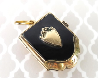 May include: A gold-toned pendant with a black enamel surface and a shield-shaped gold emblem. The pendant has a rectangular shape with rounded edges and a decorative gold border. A small gold loop is at the top for a chain.