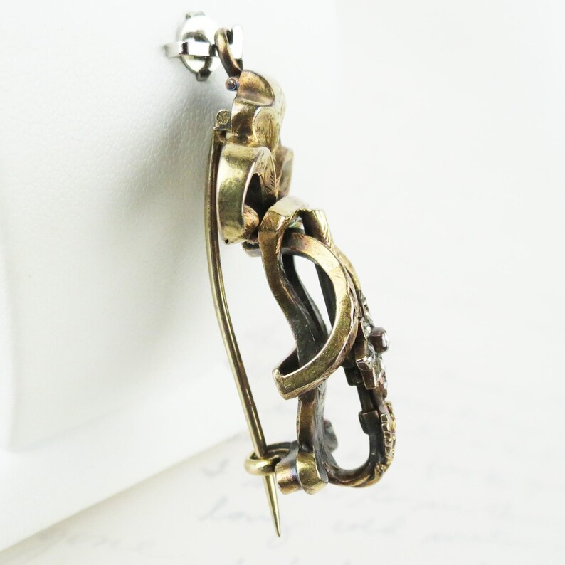 May include: Vintage gold-toned brooch with intricate looping design and small, embedded gemstones. The pin features a classic clasp and a decorative, ornate style, suitable for formal wear or adding a touch of elegance.