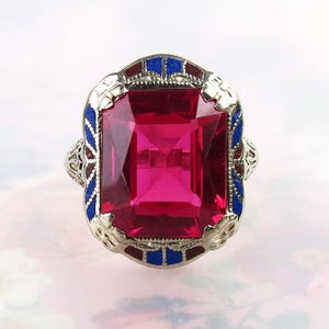 May include: A silver-toned ring featuring a large, rectangular, faceted deep pink stone at the center. The metal setting is decorated with intricate filigree patterns and accented with small, vibrant blue and dark red enamel inlay details.