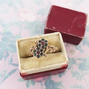 May include: A vintage gold ring with two floral clusters, each with dark blue and red gemstones. The ring is displayed in a velvet-lined box with a red exterior. The ring's design features intricate scrollwork.