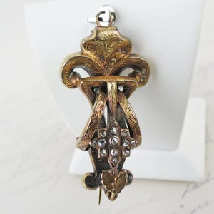 May include: A gold-toned metal brooch featuring an ornate, engraved scrollwork design with a central cross-shaped element set with small, clear, faceted stones. The piece includes a top loop and a pointed bottom with leaf-like detailing.