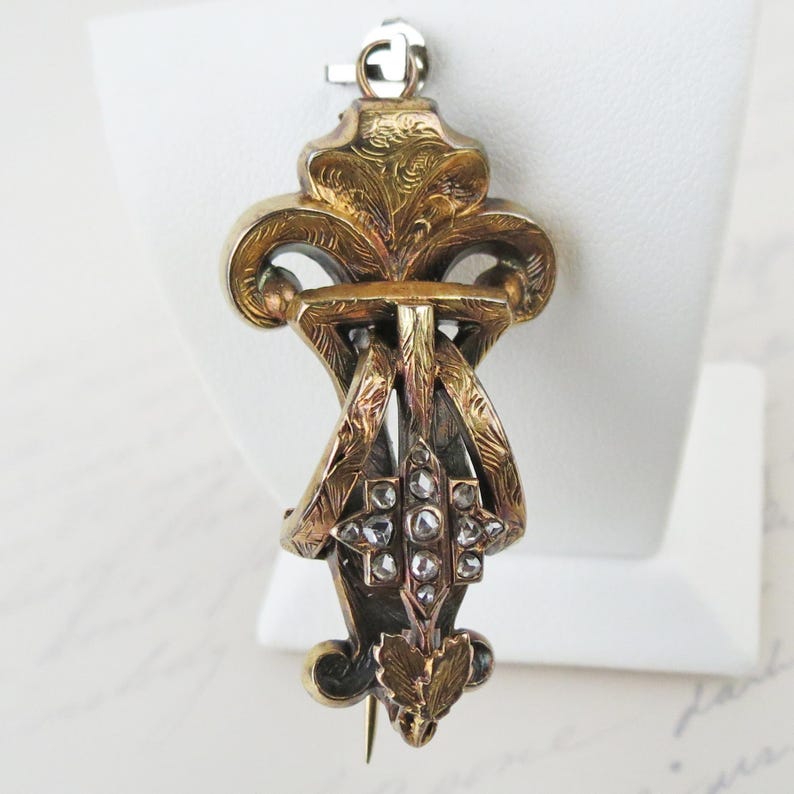 May include: A gold-toned metal brooch featuring an ornate, engraved scrollwork design with a central cross-shaped element set with small, clear, faceted stones. The piece includes a top loop and a pointed bottom with leaf-like detailing.