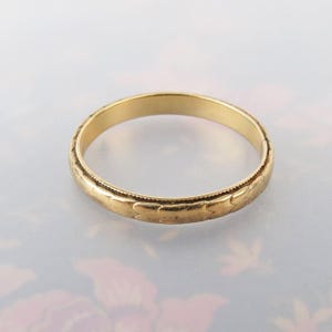 May include: A thin gold band ring featuring a repeating engraved pattern of small, stylized leaf or petal shapes along the outer circumference, bordered by a fine milgrain texture on the top and bottom edges of the metal surface.