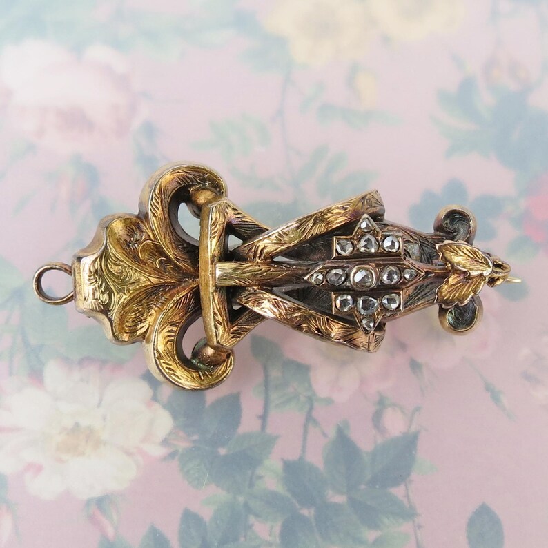 May include: An antique gold-toned brooch with intricate floral and geometric designs. The brooch features a central diamond-studded section and ornate detailing, showcasing a vintage aesthetic. The piece has a pin closure and a loop for attachment.