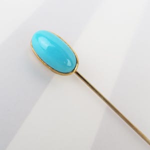 May include: A stick pin featuring a smooth, oval-shaped, bright turquoise-colored stone set in a polished gold-toned metal bezel. The pin has a long, slender, straight gold-toned metal shaft extending from the base of the stone setting.