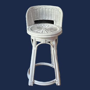 May include: White wicker bar stool with a round seat and backrest. The seat and back are woven with a circular pattern. The stool has a footrest and four legs. The chair is painted white and is set against a dark blue background.