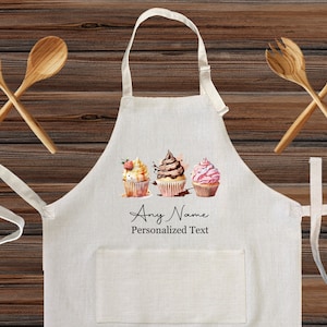 May include: A cream-colored apron with a pocket and a watercolor design of three cupcakes. The apron has the text "Any Name Personalized Text". Two wooden spoons are crossed above the apron.