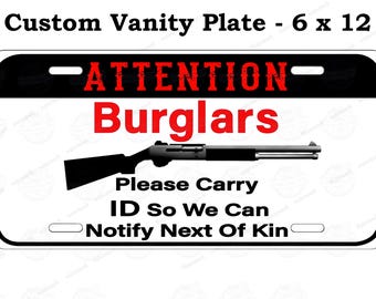 Può includere: Una targa personalizzata bianca con bordi neri e la scritta "ATTENTION Burglars" in rosso. Una grafica di fucile nero è centrata sotto. La targa misura 15 x 30 cm.