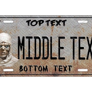May include: A rusty metal license plate with a mummy design. The plate has the text "TOP TEXT", "MIDDLE TEXT", and "BOTTOM TEXT" on it. The plate also has the text "OCT" and "98" on it.