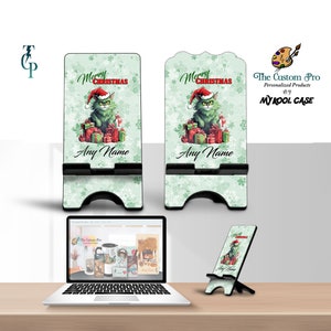 May include: A green and white phone stand with a Christmas cat design. The stand has a cutout for a phone and a kickstand. The text "Merry Christmas" is written on the stand. The text "Any Name" is written on the stand.