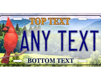 Può includere: Targa con sfondo montuoso e un cardinale rosso acceso appollaiato su un ramo. La targa ha campi di testo: "TOP TEXT", "ANY TEXT" e "BOTTOM TEXT".