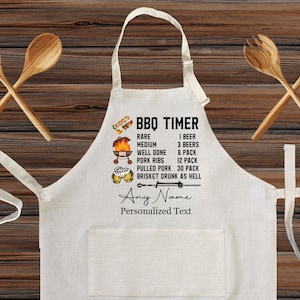 May include: A white cotton BBQ apron with a black graphic that says "BBQ Timer" and lists different levels of doneness for meat with corresponding beer amounts. The graphic also includes a grill, beer mugs, and a sword. The apron has a pocket and adjustable neck straps. The text "Any Name" and "Personalized Text" is also included.