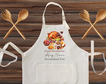 Può includere: Un grembiule bianco con una tasca, con un'illustrazione della cena del Ringraziamento e il testo "Happy Thanksgiving" e "Any Name Personalized Text". Due cucchiai di legno sono incrociati sopra il grembiule.