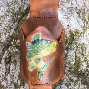 May include: A brown leather pouch with a tooled design of a fish. The pouch has a loop at the top for hanging.