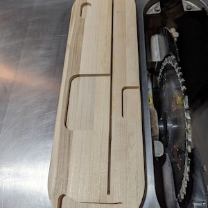 Puede incluir: Una tabla de madera con recortes para sujetar herramientas. La tabla es de color marr&oacute;n claro y tiene un acabado liso. Est&aacute; sentada en una sierra de mesa.