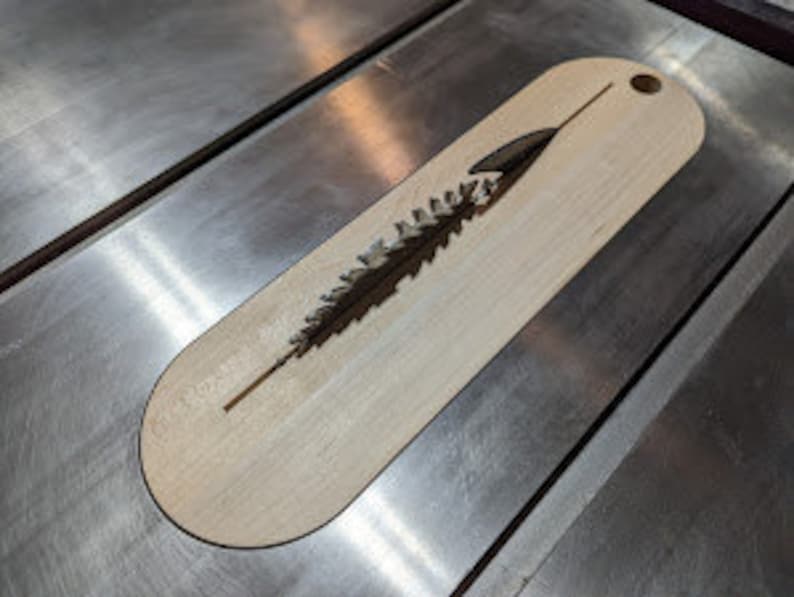 Puede incluir: Un marcap&aacute;ginas de madera con un dise&ntilde;o de pino cortado con l&aacute;ser. El marcap&aacute;ginas tiene forma ovalada y tiene un agujero en la parte superior para una cinta.