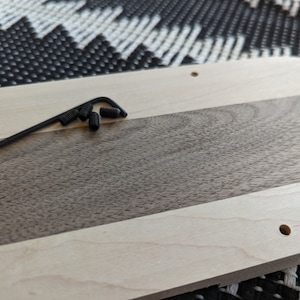 Puede incluir: Un primer plano de una tabla de cortar de madera con una parte superior de madera clara y una parte inferior de madera oscura. La tabla tiene un agujero en la esquina superior derecha y una peque&ntilde;a herramienta de metal con tornillos negros est&aacute; sobre la tabla.