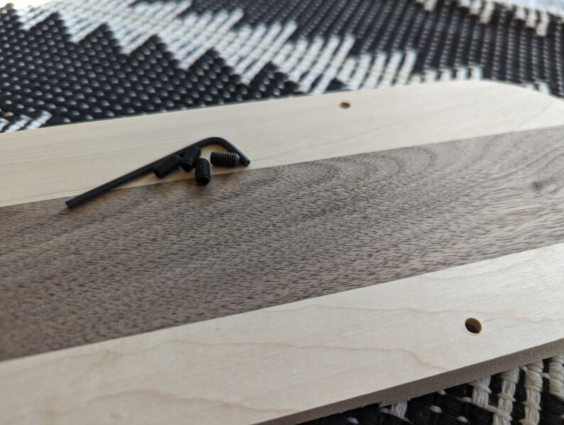 Puede incluir: Un primer plano de una tabla de cortar de madera con una parte superior de madera clara y una parte inferior de madera oscura. La tabla tiene un agujero en la esquina superior derecha y una peque&ntilde;a herramienta de metal con tornillos negros est&aacute; sobre la tabla.