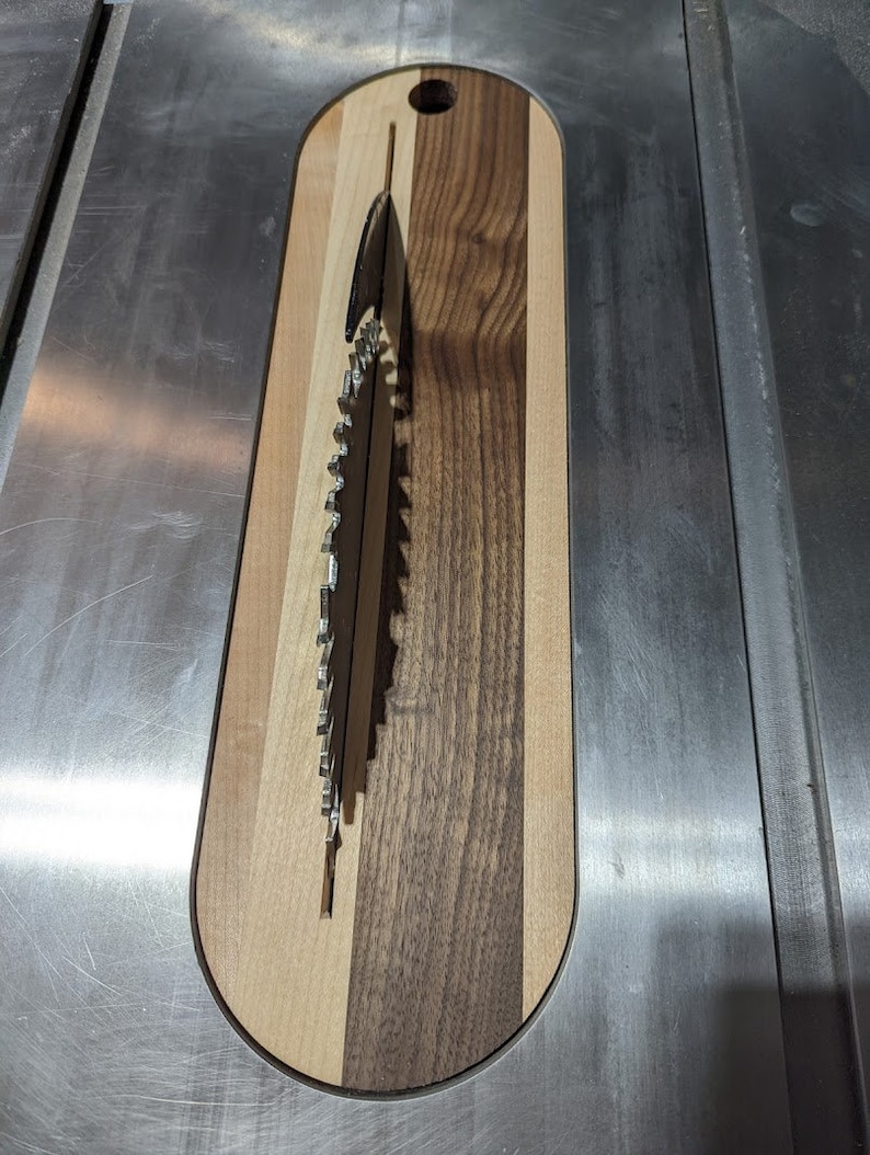 Puede incluir: Una tabla de cortar de madera con un recorte circular para una hoja de sierra. La tabla est&aacute; hecha de tres tipos diferentes de madera: arce, nogal y cerezo. La hoja de sierra es visible a trav&eacute;s del recorte.